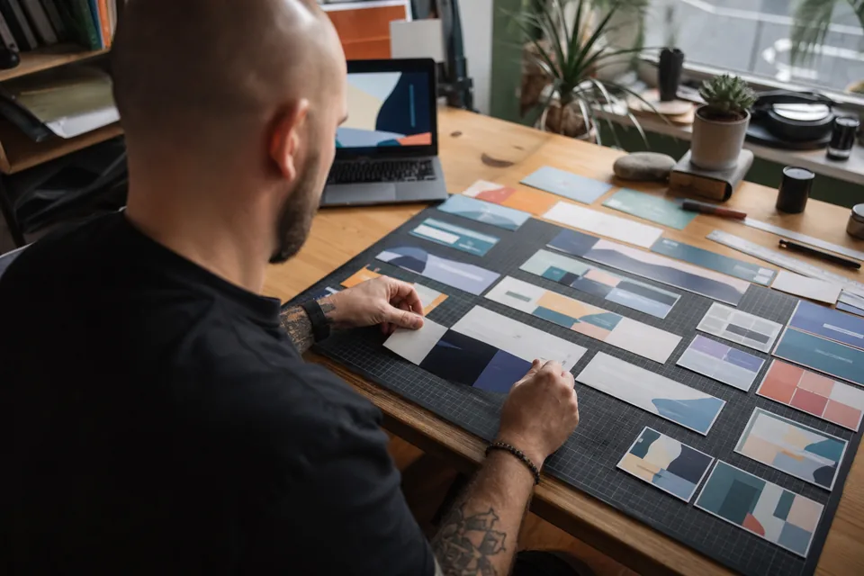 Web designer at a desk sorting printed landing page sections into rows of heroes, pricing tables and footers