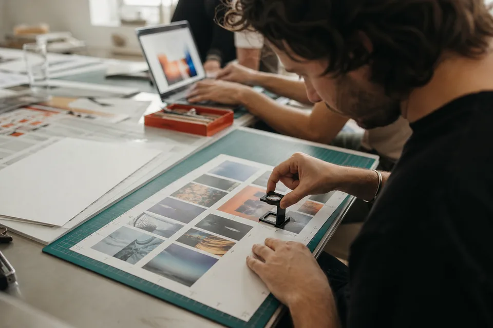 Hands with a cutting mat, crop marks on printed layouts and a loupe on a contact sheet